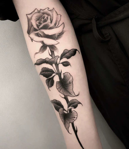 Vertical black-and-grey rose stem temporary tattoo with bold leaf shading on forearm
